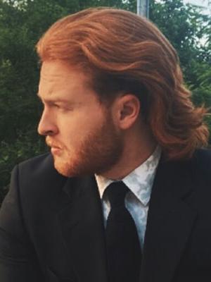 White man with red hair and a red beard wearing a black suit
